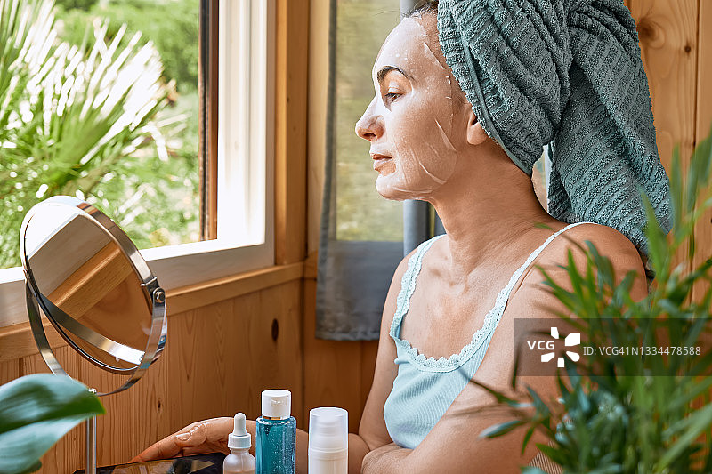 护肤习惯。脸上敷着保湿面膜的中年妇女望着窗外。提升眼下抗皱面膜。胶原蛋白面膜，护肤和水疗概念。图片素材