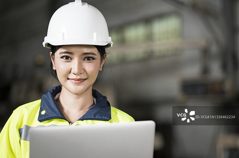 女机械工程师在工厂里用笔记本电脑工作，戴着白色安全帽，穿着安全背心。擅长机器人焊接工作过程的研究、设计、开发、改进。图片素材