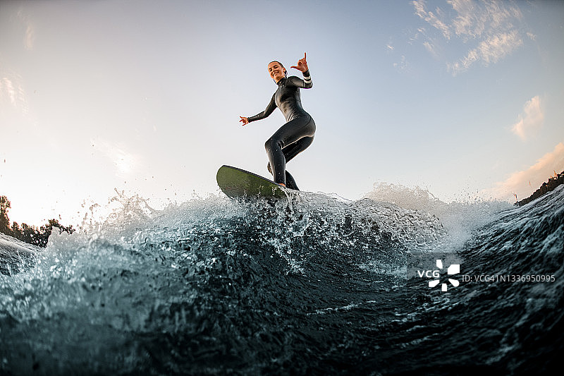 快乐的女人穿着潜水服在冲浪板上冲浪，顺流而下，挥挥手图片素材