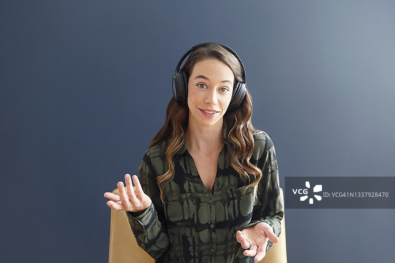 一位留着棕色长发的千禧一代白人女性，穿着休闲装，戴着无线耳机坐在蓝色墙壁前进行视频会议。图片素材