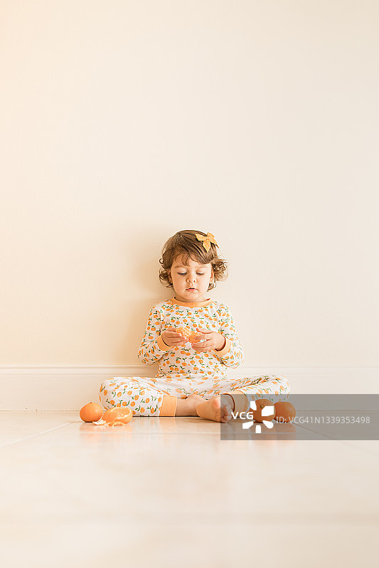 2021年9月，两岁的小女孩坐在地板上玩和吃橘子，穿着橙色睡衣和橙色的头发蝴蝶结在家里图片素材