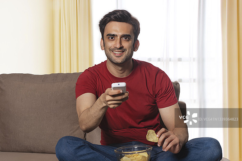 一个快乐的男人拿着遥控器坐在沙发上吃薯片在他的房间。图片素材