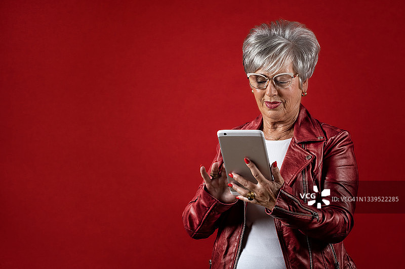 年长的白发女人戴着眼镜，在红色背景前使用数码平板电脑图片素材