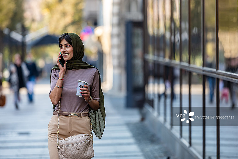美丽的穆斯林中年妇女在街上一边打电话一边喝咖啡图片素材