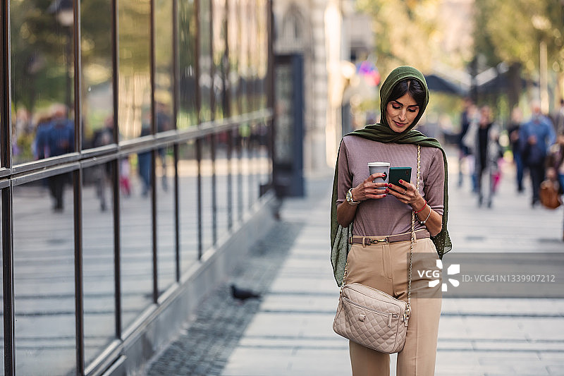 一名中年穆斯林女性拿着一杯咖啡，在智能手机上打字。图片素材