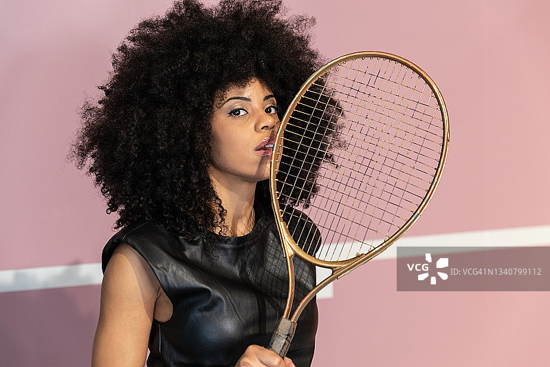 非洲裔美国女模特，一头非洲式的短发，穿着时尚的服装，站在粉红色背景的摄影棚，拿着网球拍看着镜头图片素材