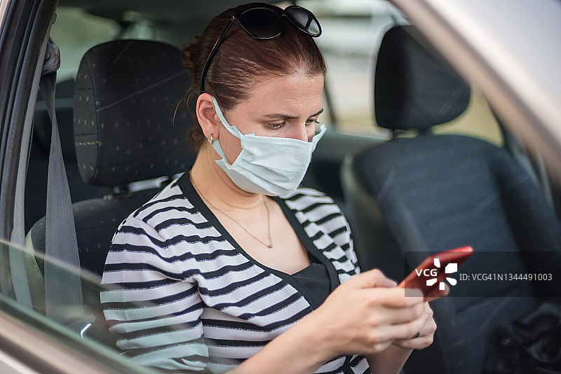 年轻女子在车里戴着保护面罩用智能手机。图片素材