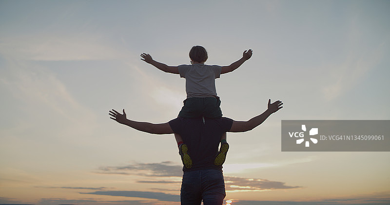 爸爸把儿子抱在肩上。他们张开双臂，在日落的天空下享受自由图片素材