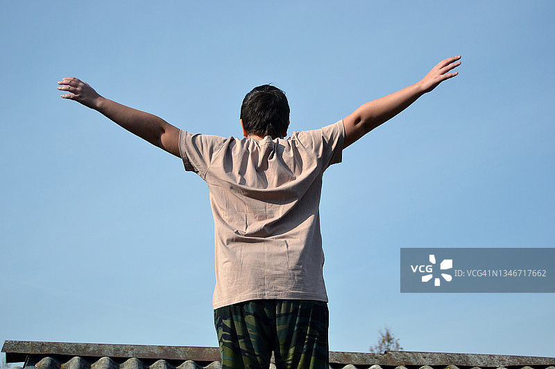 一个十几岁的男孩站在屋顶上，在蓝天的映衬下。图片素材