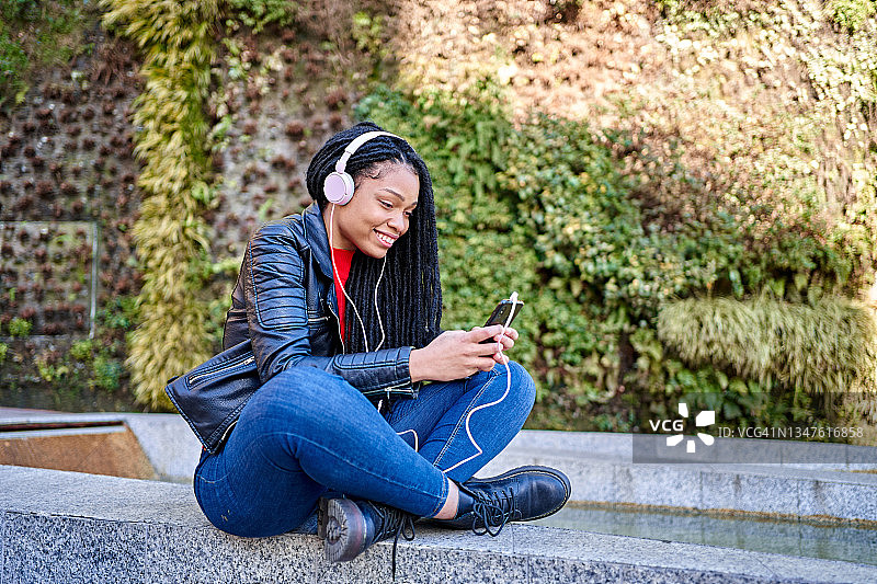 图为一名微笑着拿着手机的非裔美国妇女在城市的街道上聊天或听音乐。头发被编成了脏辫。图片素材