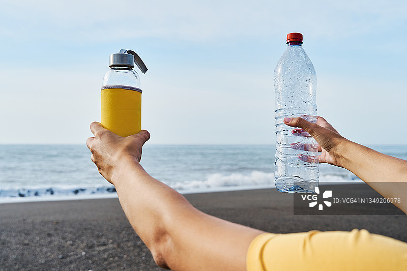一个女人在海边用手比比一次性塑料瓶和可重复使用的玻璃瓶。图片素材