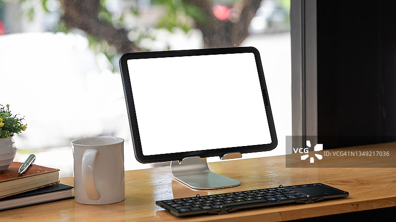 工作空间的数字平板电脑空白的白色屏幕与咖啡杯键盘放在木桌。模拟。图片素材
