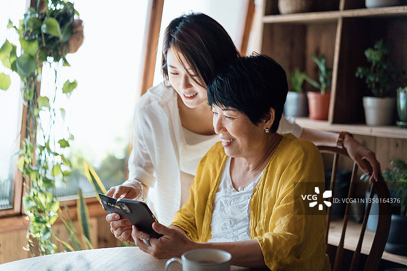 快乐的亚洲资深妈妈和女儿一起用智能手机网购，在家用信用卡付款图片素材
