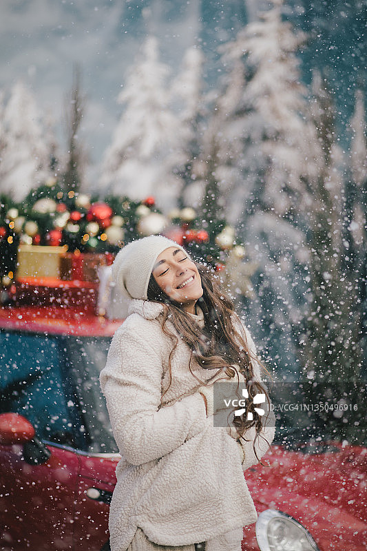 快乐的美丽的女人穿着针织的毛衣和羊毛帽站在红色的汽车旁边，顶上有圣诞树在下雪。图片素材
