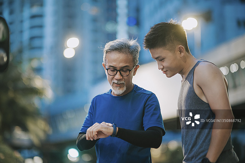 一位亚裔中国老人和他的儿子在深夜跑步后用健身追踪器检查心跳图片素材