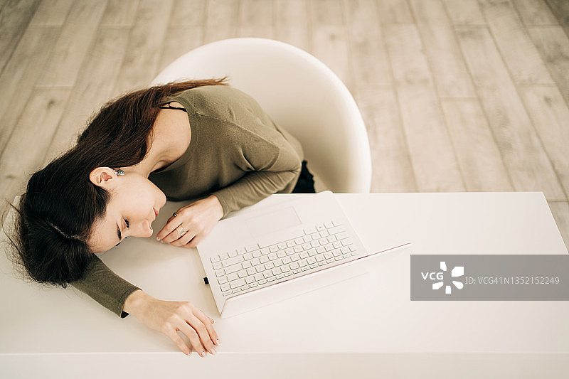 一个棕色长发的年轻漂亮女人闭着眼睛，躺在桌子上睡着了。她在家里用笔记本电脑工作，很累。远程工作的概念图片素材