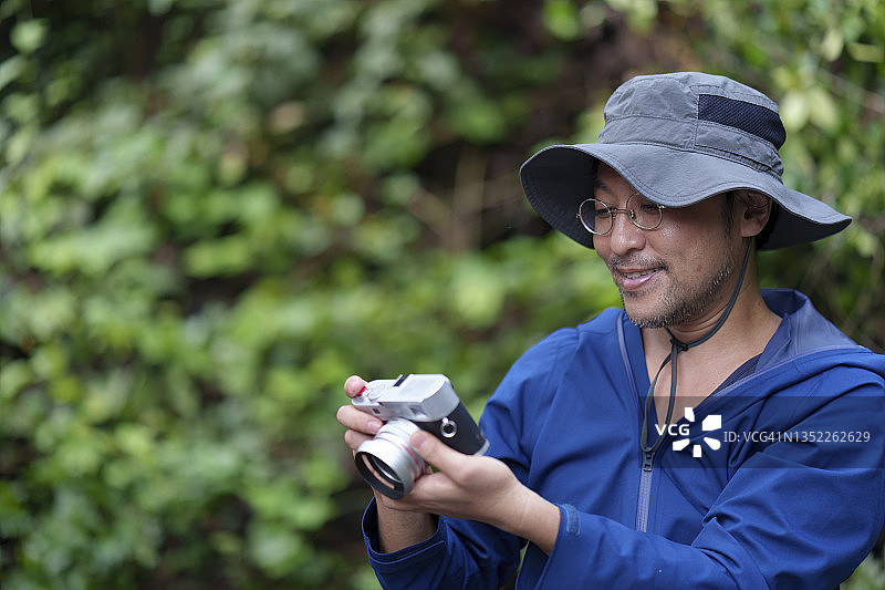 一名日本男子在热带森林里拿着相机看着照片，享受着假期。图片素材