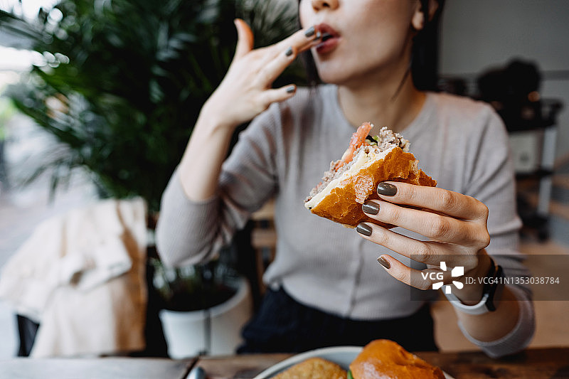 剪辑，年轻的亚洲女人在咖啡馆吃着新鲜的美味芝士汉堡，舔着她的手指。享受她的午餐!人与食物的概念图片素材