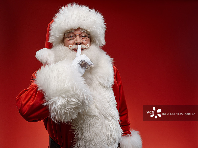 有趣的圣诞老人展示了一个秘密的标志，他的手指放在嘴唇上，站在一个红色的背景，在一个专业的照明工作室图片素材