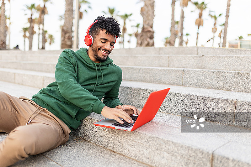 开朗的西班牙裔学生男子坐在户外和工作的笔记本电脑，同时听音乐图片素材