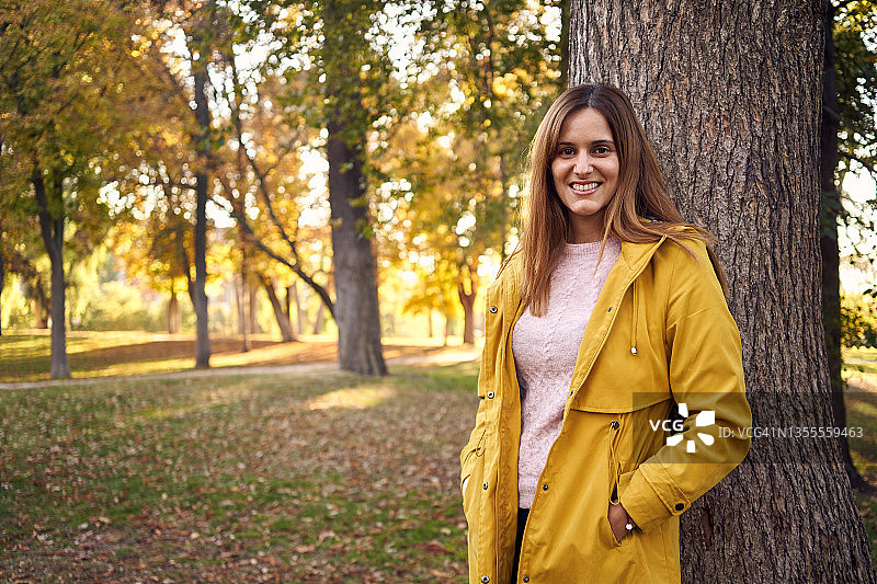 穿着便服和黄大衣的年轻女子倚在树干上，在秋天的公园与复制空间。休闲、自然、生活理念。图片素材