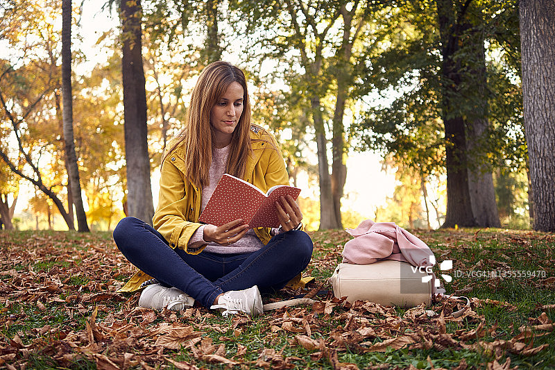 穿着便服和黄大衣的年轻女子在树木环绕的公园里坐着看书。24、闲暇观，自然观，读书观。图片素材