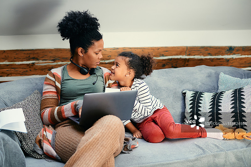 一个非洲裔美国小女孩正在和她的妈妈说话，她的妈妈正在家里用笔记本电脑工作。图片素材