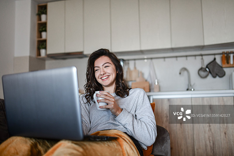 美丽的女性躺在厨房沙发上喝咖啡和看有趣的电影在笔记本电脑上图片素材