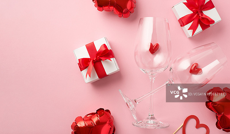 情人节装饰俯视图照片小心在两个葡萄酒杯白色礼品盒与红色丝带蝴蝶结和心形气球在孤立的淡粉色背景图片素材