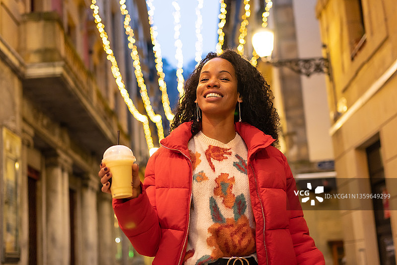 刻画黑人少数民族妇女冬天在城市的夜晚。拿着外卖橙汁，穿着红色夹克微笑着走在灯光明亮的街道上图片素材