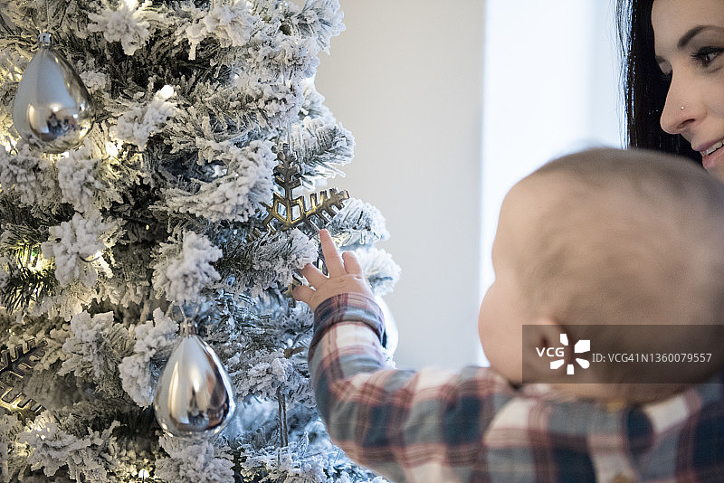 在英国苏格兰边境梅尔罗斯的一所房子里，一位年轻的母亲抱着她的孩子微笑着，她的女儿正伸出手去拿挂在窗户边的白色圣诞树上的装饰品图片素材