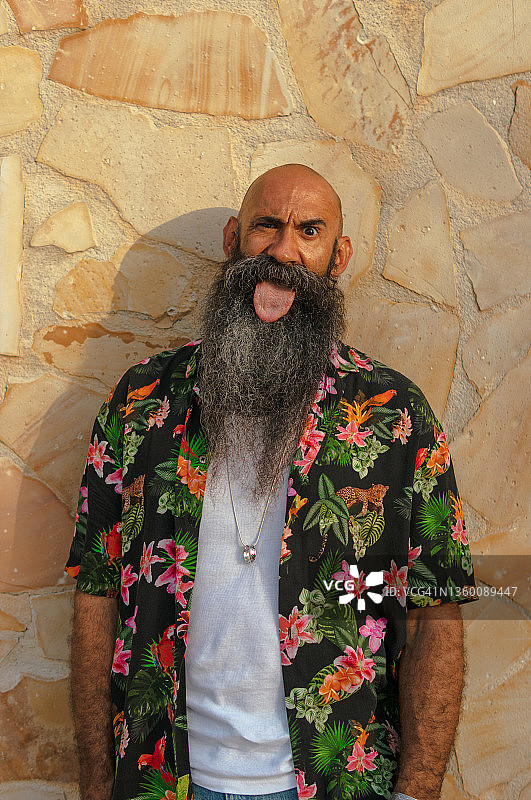 穿着夏威夷衬衫，露出舌头的光头大胡子男子。图片素材