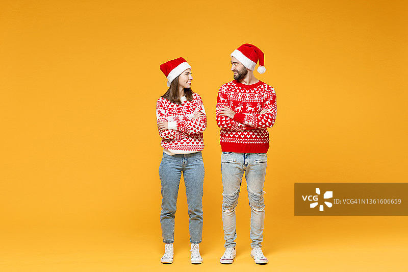 整个长度微笑的年轻的圣诞老人夫妇朋友，男人，女人在毛衣圣诞帽，手交叉看着对方孤立在黄色的背景。祝新年欢度佳节理念。图片素材