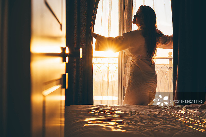 清晨，女人打开窗帘从大窗户欣赏景色图片素材