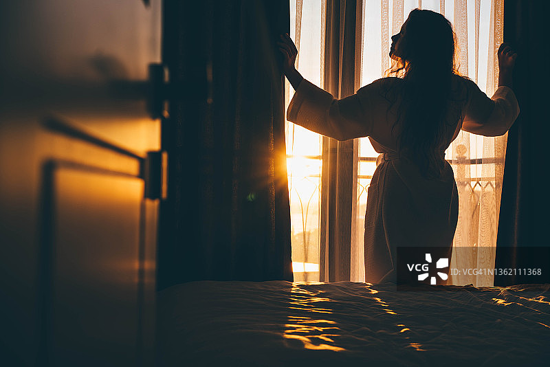 清晨，女人打开窗帘从大窗户欣赏景色图片素材