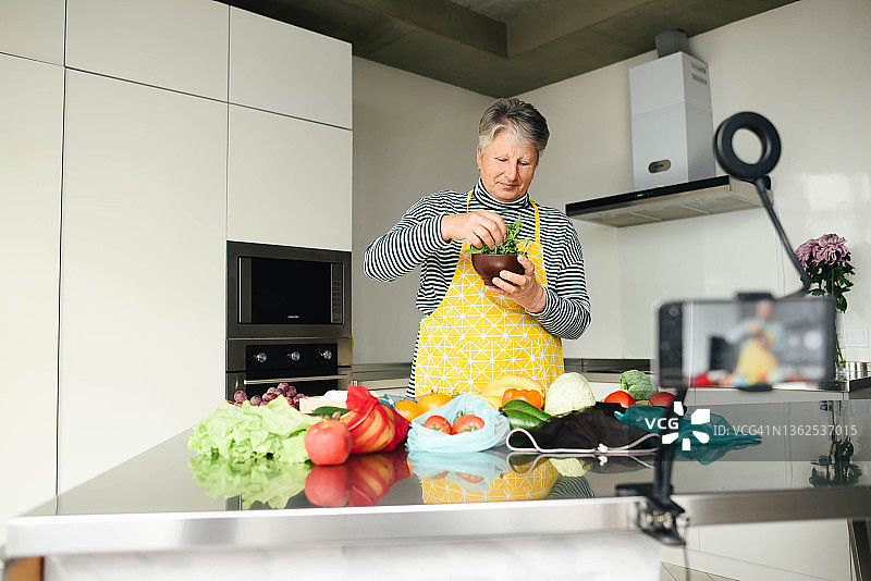 一位上了年纪的烹饪博客作者正在厨房里拍摄视频。现代的美食。天然的蔬菜。健康的生活方式。环保的食物。一位上了年纪的女人。黄色的围裙。美食博客。图片素材