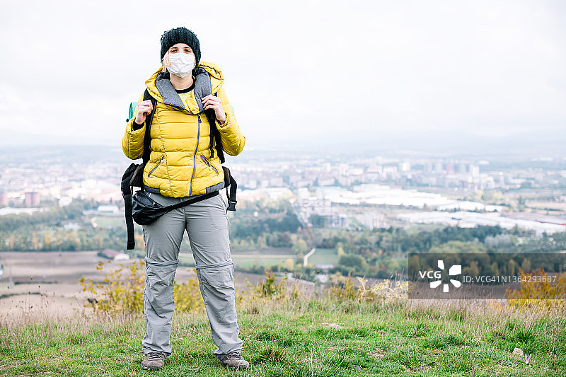 一个带着面具的女背包客站在山上，背后是城市，看着镜头图片素材