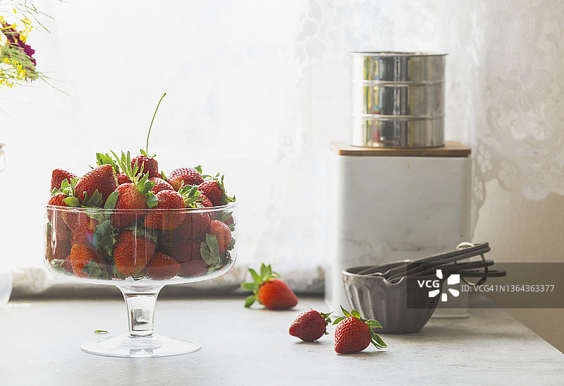 玻璃碗装满了新鲜的草莓，量杯和储存容器在厨房桌子上的窗户背景与自然光线图片素材