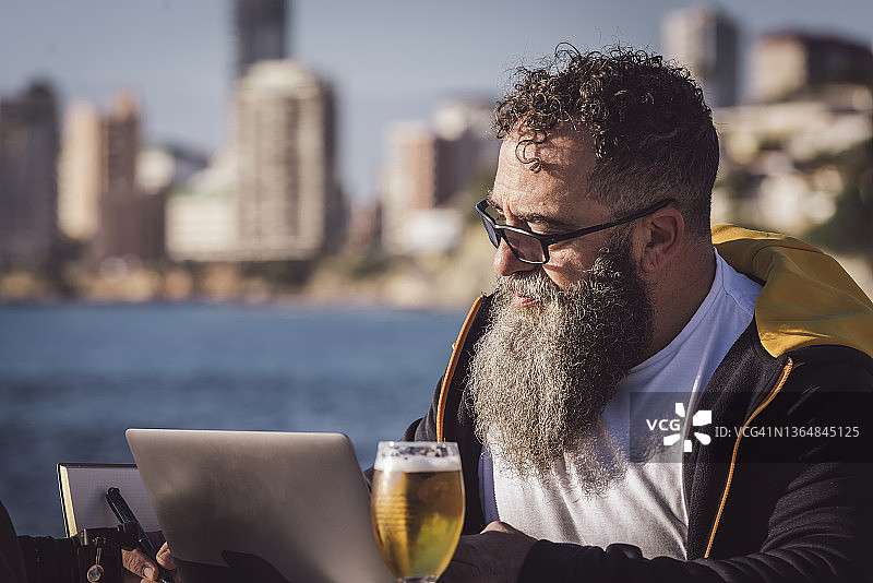 留着长胡子的Homabre坐在海边的一家咖啡馆里，一边用他的笔记本电脑在线工作。图片素材