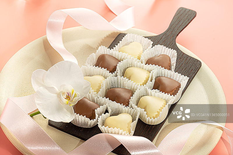 白色兰花花和白色和黑色巧克力在心形的盘子和粉红色的丝带在柔和的桃红色背景。图片素材