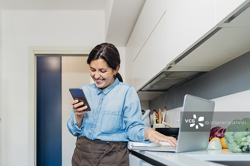 微笑的女人用手机和笔记本电脑站在厨房柜台在家里图片素材