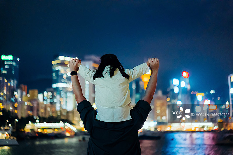 年轻慈祥的亚洲父亲肩上扛着小女儿，在夜晚俯瞰着维多利亚港的海滨。享受父女相处的时光。亲情与生活方式图片素材