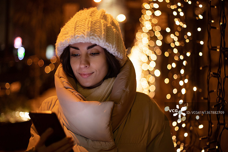 美丽微笑的女人在白色冬季帽子和夹克的特写肖像，而她持有和使用她的智能手机在户外行走与城市圣诞照明的背景图片素材