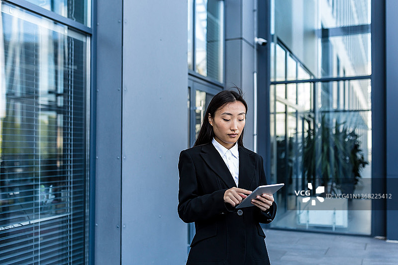 亚洲女商人在商务穿着西装站着工作，手拿平板电脑的背景现代办公大楼中心外面。男性使用智能手机，使用手机户外城市街道图片素材