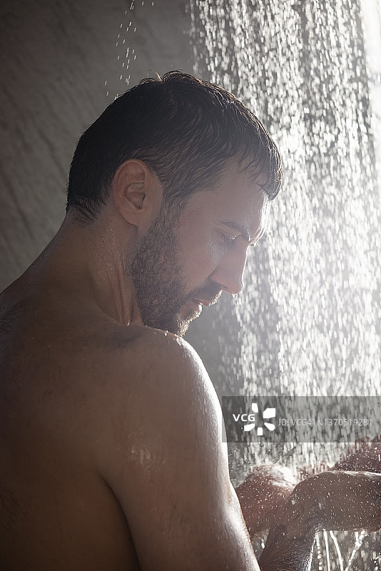 英俊的年轻人站在滴水下洗澡，在家里的现代浴室里清洗身体和头部。Selfcare和健康图片素材