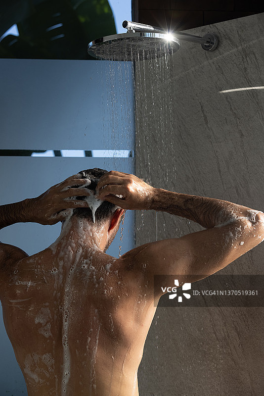 一个男人在家里的现代浴室里洗澡，洗头发和身体，站在滴水下。Selfcare和健康图片素材