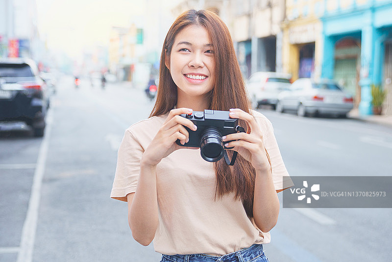 年轻的女游客用老式相机在城市里开心地微笑着。快乐的年轻女人，带着城市地图，背着背包旅行图片素材