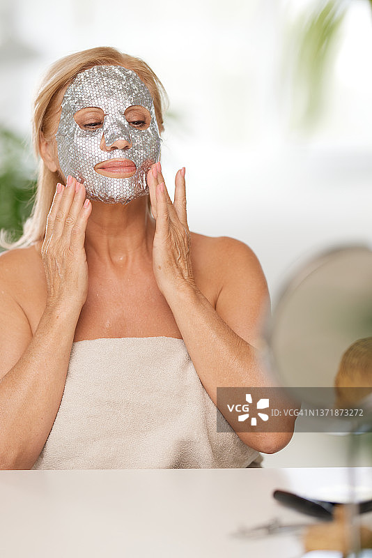 一位老妇人裹着毛巾坐在家里，把美容面膜敷在脸上。高级女性使用美容产品图片素材