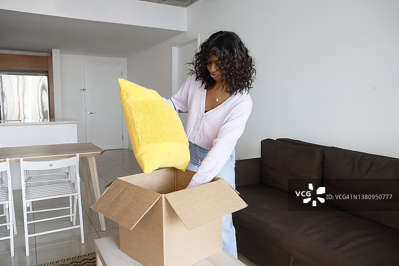 一位留着波浪短发的美国黑人Z一代女性，在她的新公寓里，穿着休闲服装，打开了一个装满家居用品的移动箱子。图片素材