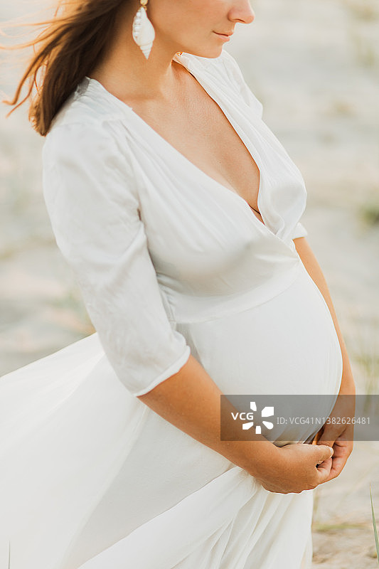 身穿白色连衣裙的孕妇抱着她的肚子。图片素材